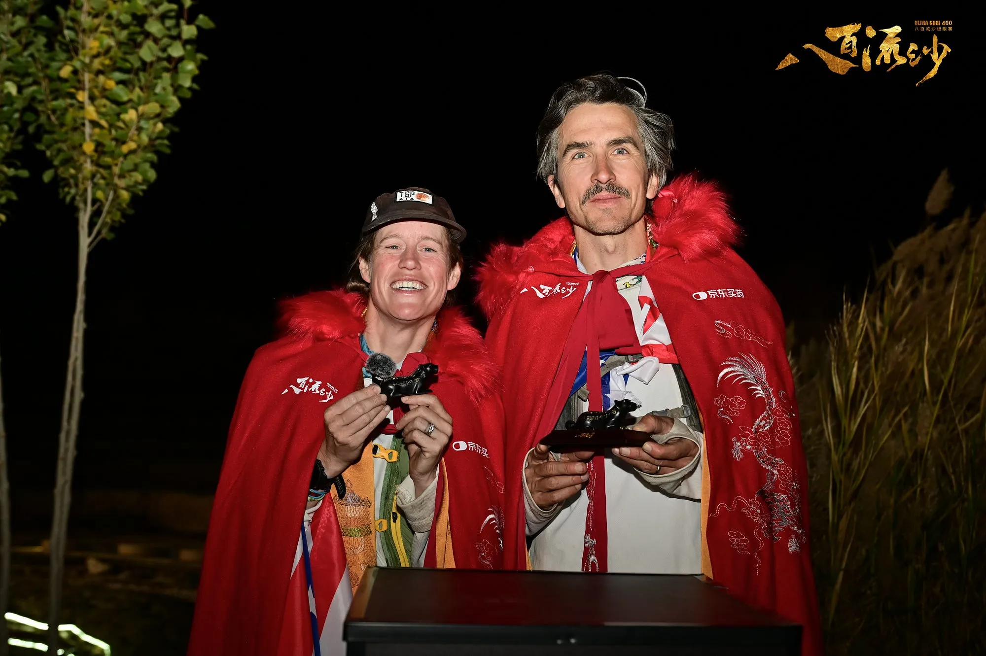 Lucy and Jack Carey at the Ultra Gobi finish at night, wrapped in red finisher capes, holding their trophies.