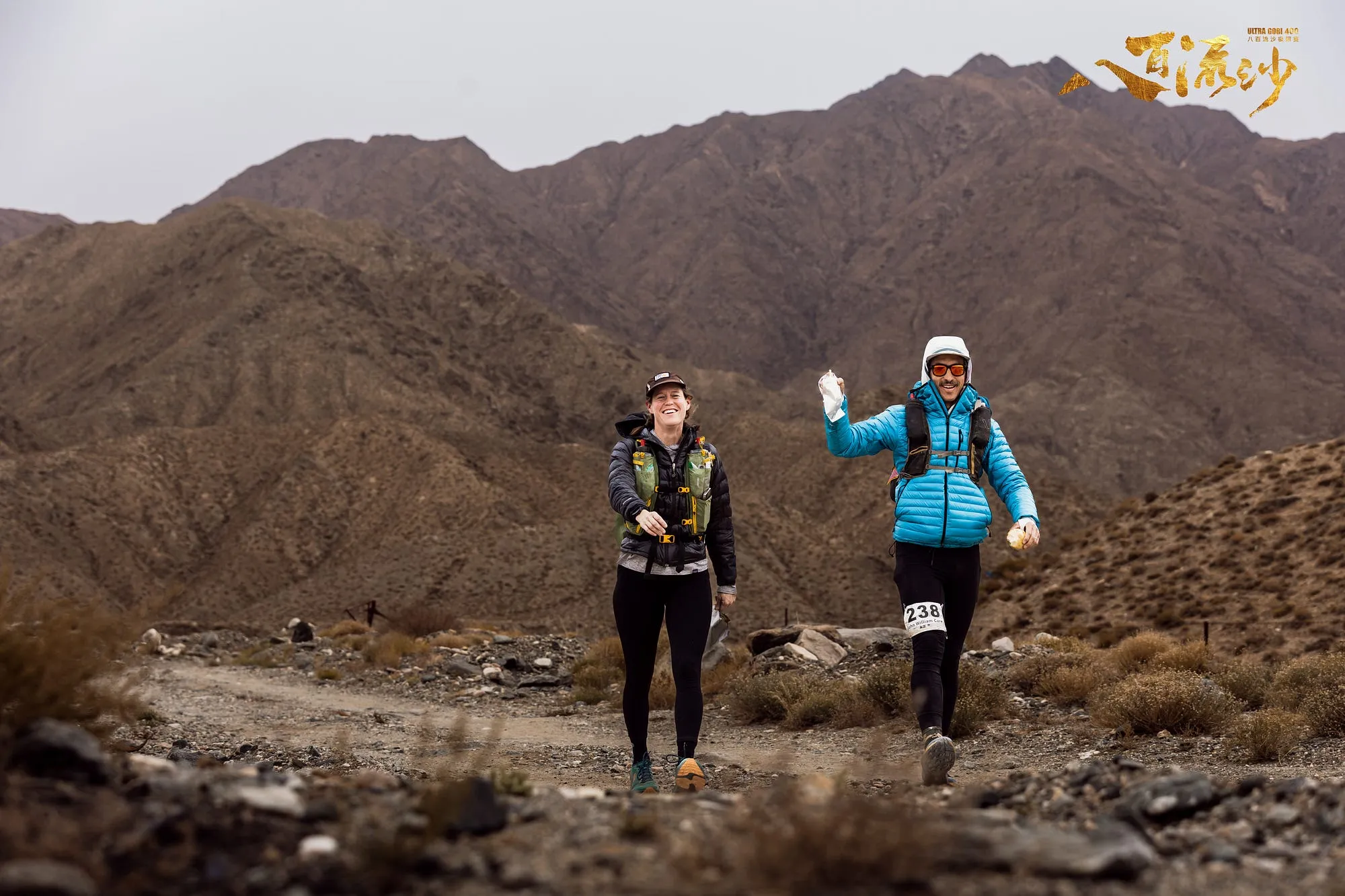Two runners moving together on a rocky trail below bare desert mountains at Ultra Gobi.