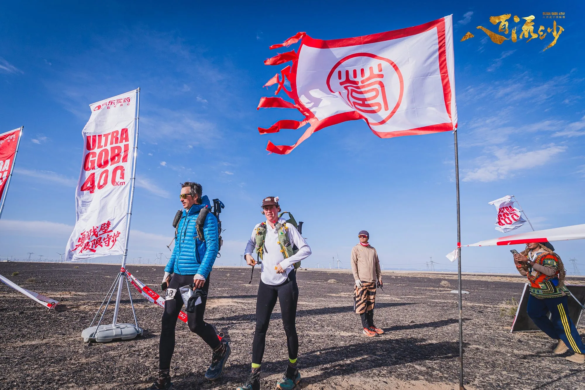 Runners at the Ultra Gobi 400K start area with red and white race flags snapping in the wind.