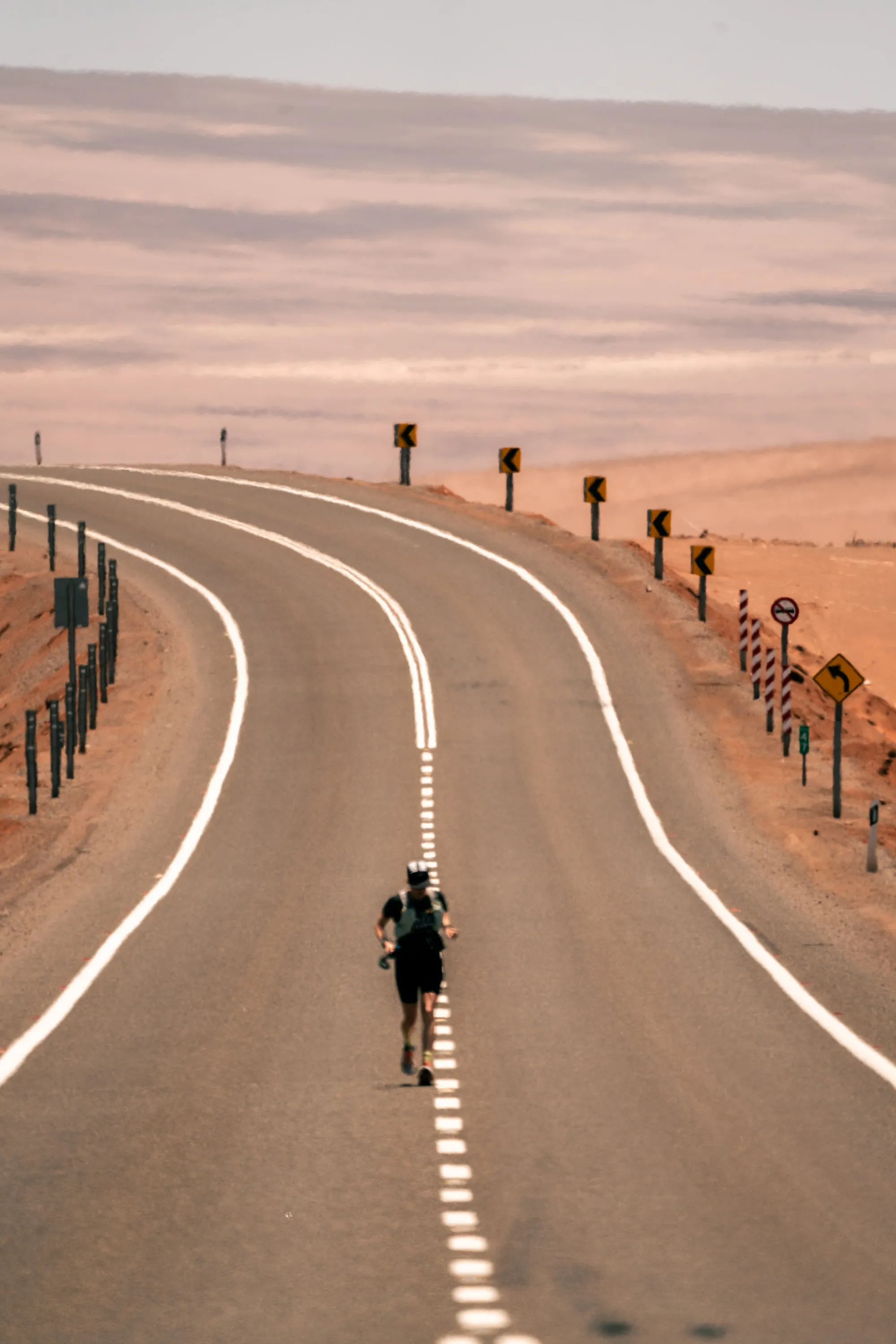 A single runner on a long curving desert highway in the Atacama at dawn, the road disappearing into pink hills.