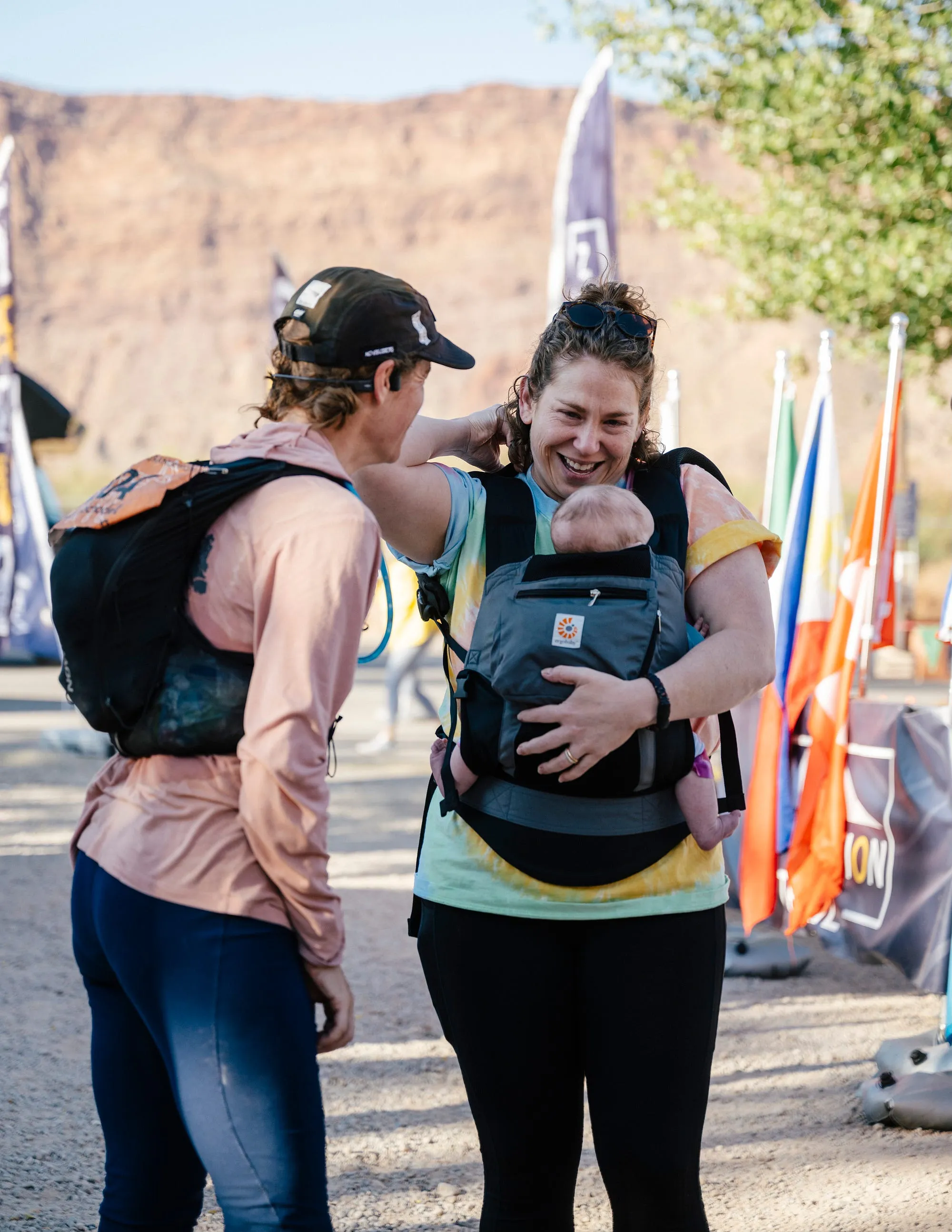 Lucy embracing a friend at the Moab 240, a baby in a carrier between them, red canyon walls behind.