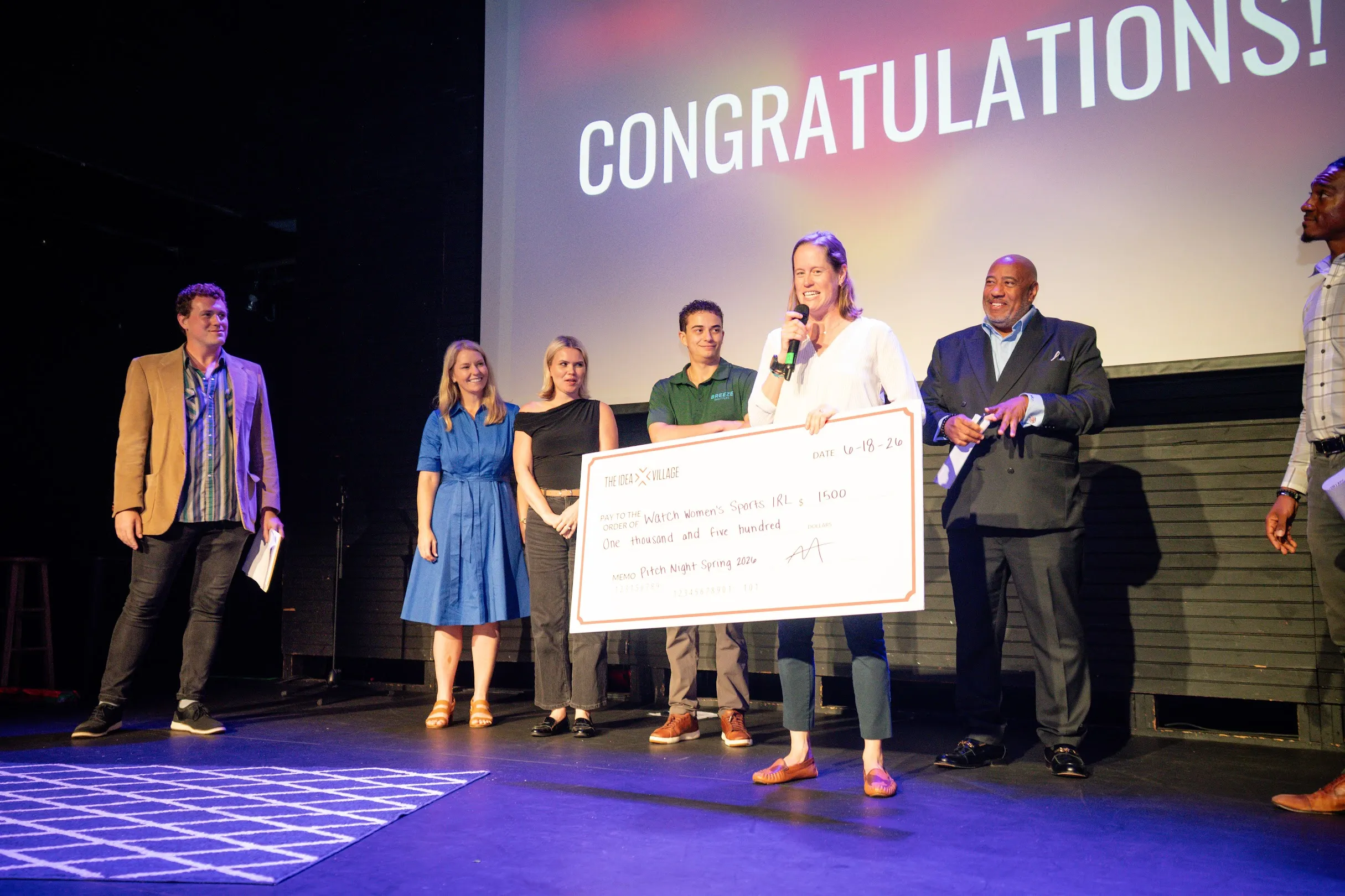 Lucy on stage at Idea Village Pitch Night holding an oversized cheque made out to Watch Women's Sports IRL for $1,500, speaking into a microphone with CONGRATULATIONS! on the screen behind her.