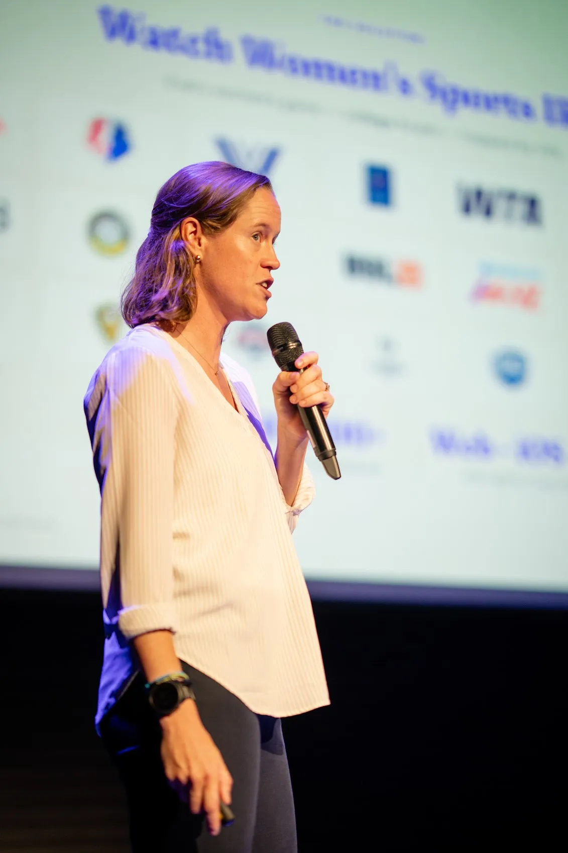 Lucy mid-pitch, microphone in hand, in front of a screen showing the Watch Women's Sports IRL slide and a wall of league logos.