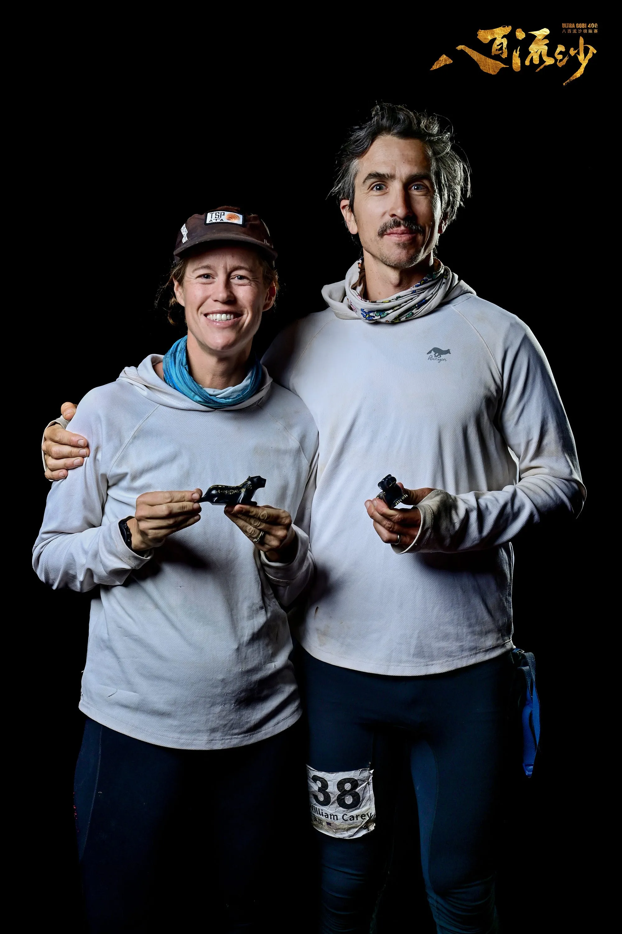 Studio-lit portrait of Lucy and Jack Carey after Ultra Gobi, arms around each other against a black background.