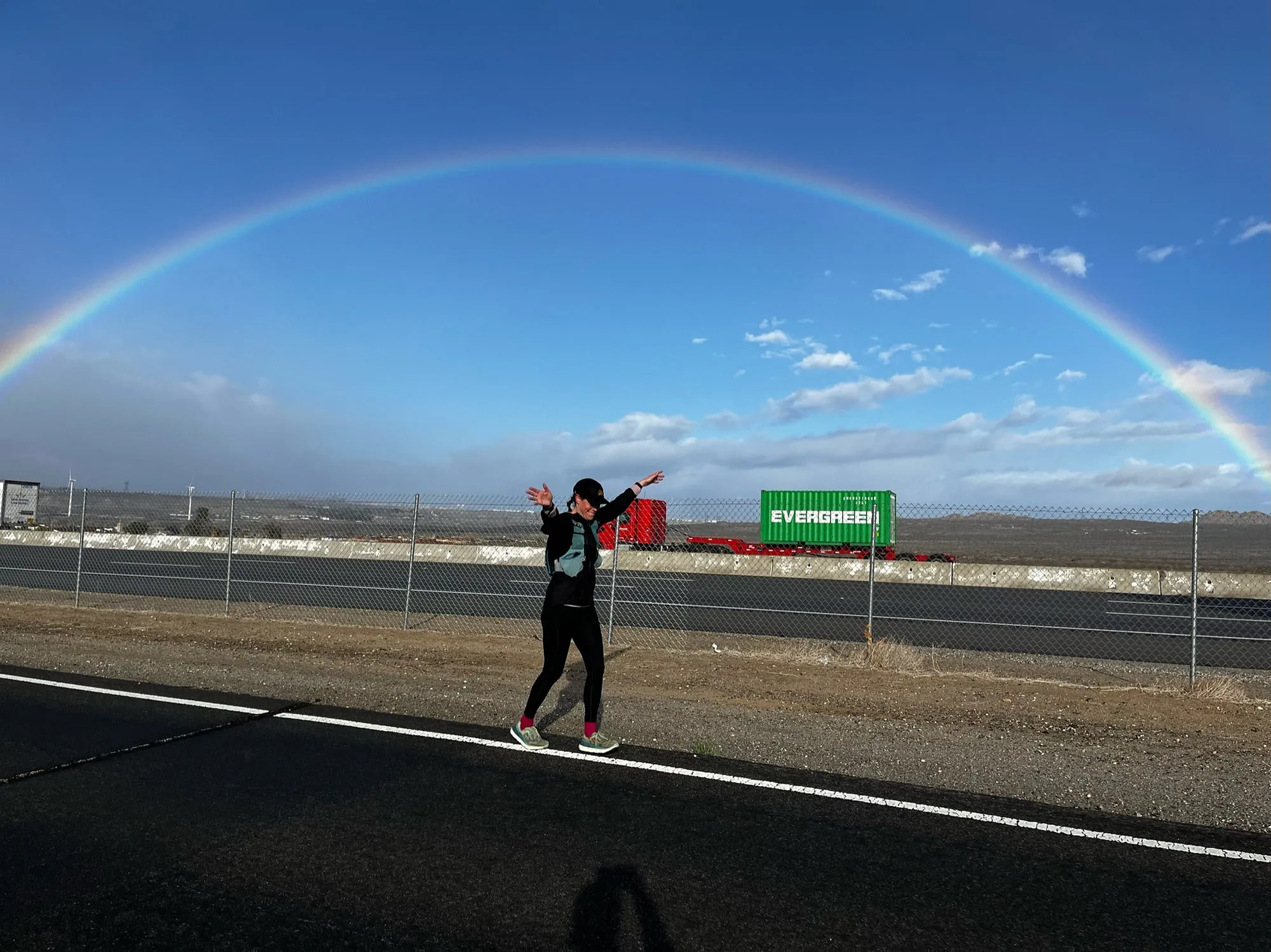 A full rainbow arcing over a desert highway with a runner small on the shoulder of the road.