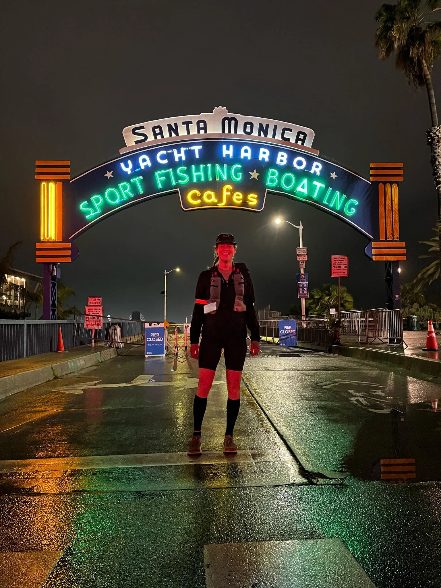 Lucy alone on wet pavement beneath the neon Santa Monica Yacht Harbor sign at night, at the start of The Speed Project.